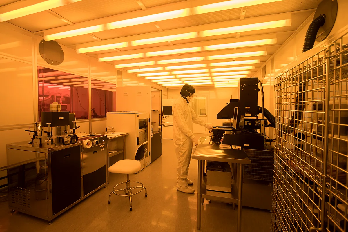 Gowned engineer in mask and gloves working at a tool in a yellow-lit lithography cleanroom